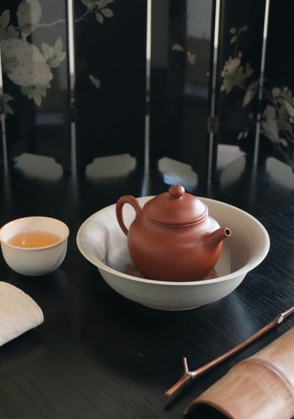 Braune Teekanne in einer hellen Schale mit einer kleinen Teetasse daneben.